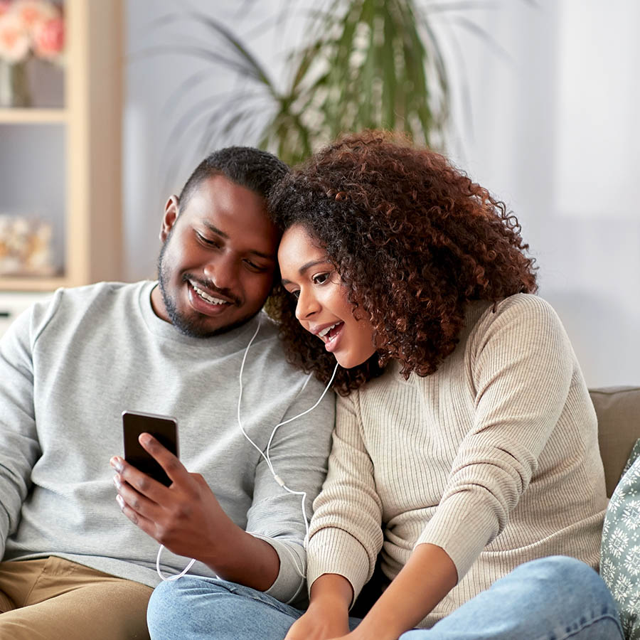 A smiling couple sits close together on a couch, sharing a pair of earphones and looking at a smartphone, enjoying music or a video in a cozy living room setting.