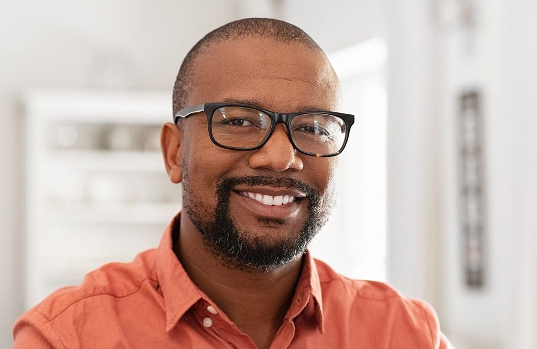 A smiling man with short hair, a trimmed beard, and glasses, wearing an orange shirt, stands indoors in a well-lit room with a blurred background.