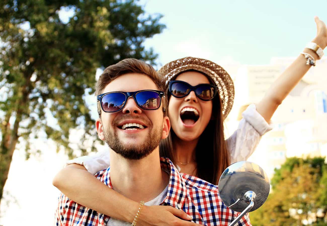 A smiling man wearing sunglasses and a plaid shirt rides a scooter with a joyful woman in sunglasses and a hat, arms raised, hugging him from behind. Trees and buildings are visible in the background.